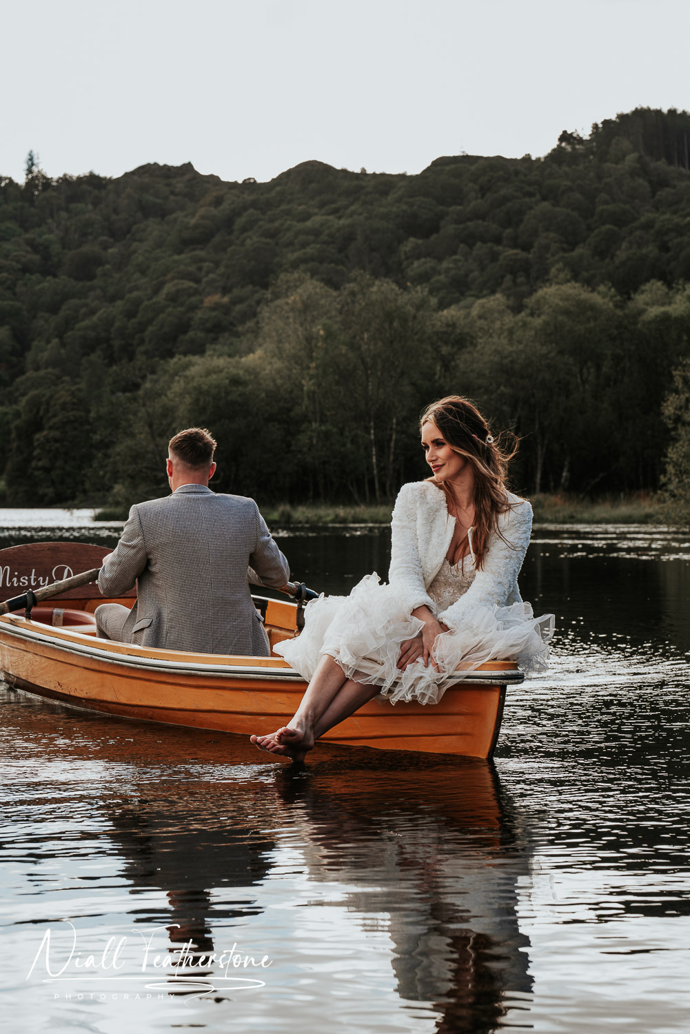 Bride Posed on Edge of rowing boat while groom rows her round the lake in Lake District